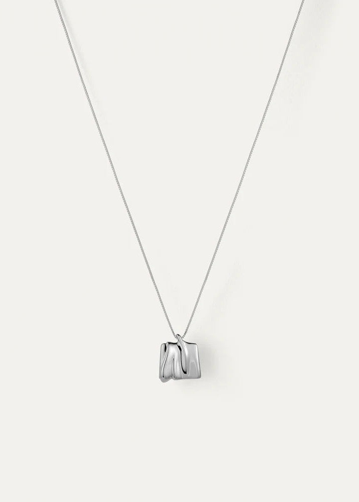 A silver pendant necklace featuring a small, sculptural geometric charm with a polished finish, suspended from a delicate silver chain against a light off-white background.