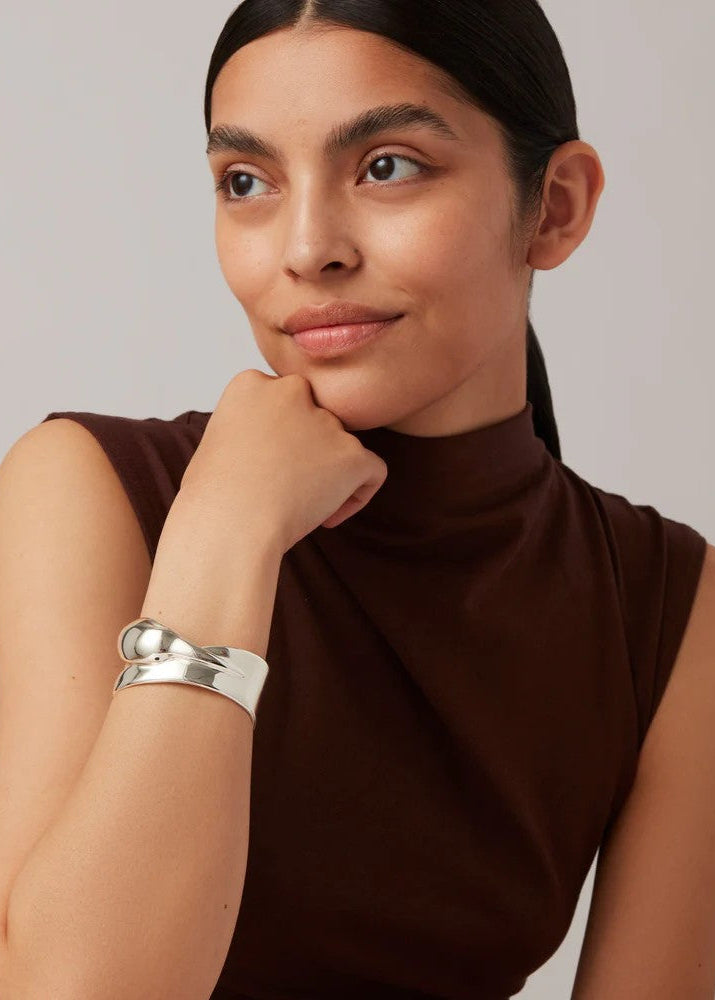 A woman wearing a wide, polished silver cuff bracelet with a knotted detail, paired with a brown sleeveless mock-neck top, photographed against a neutral light gray background.