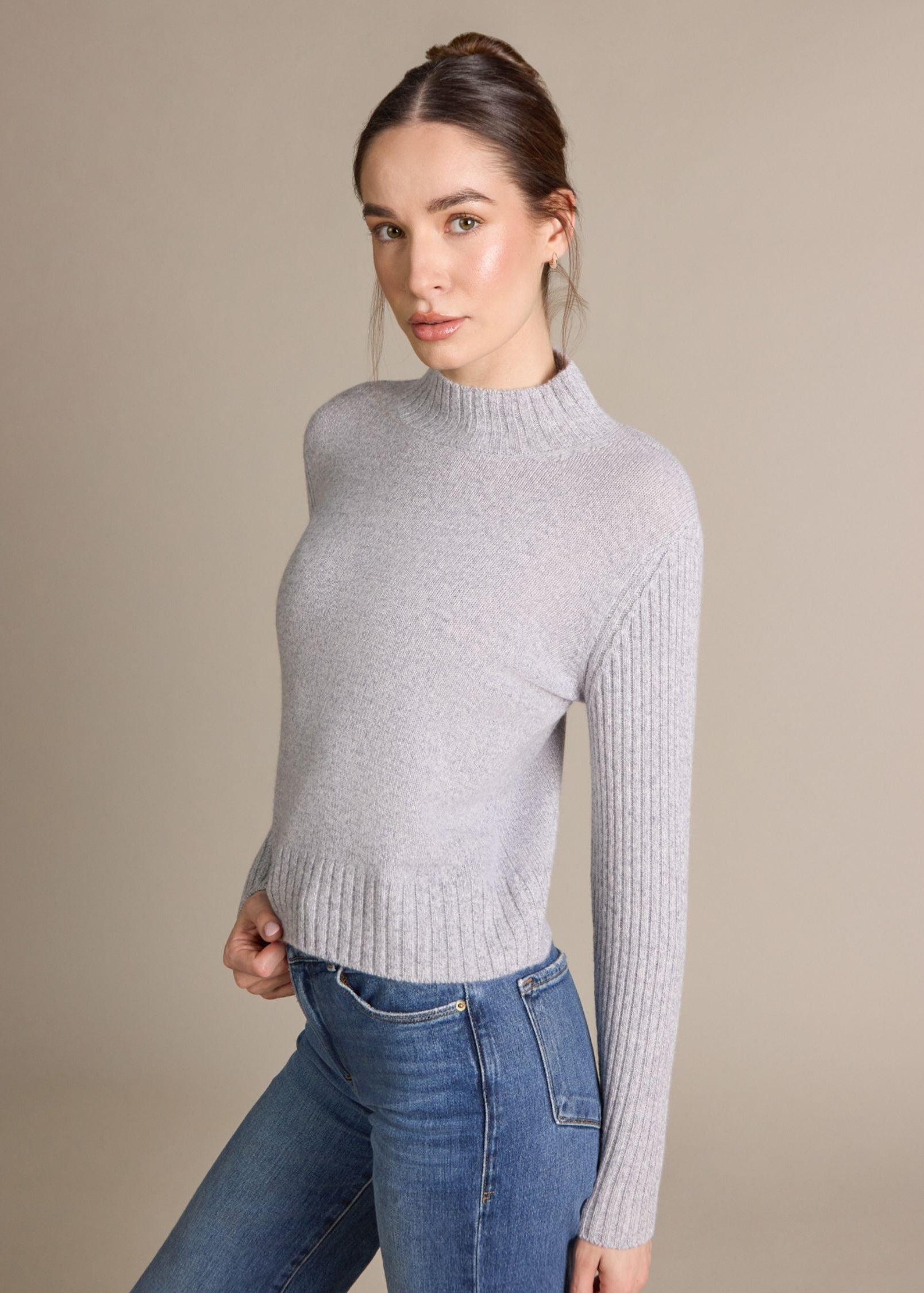 A woman modeling a light gray ribbed mock neck sweater with long sleeves, paired with blue jeans, against a solid beige studio background.