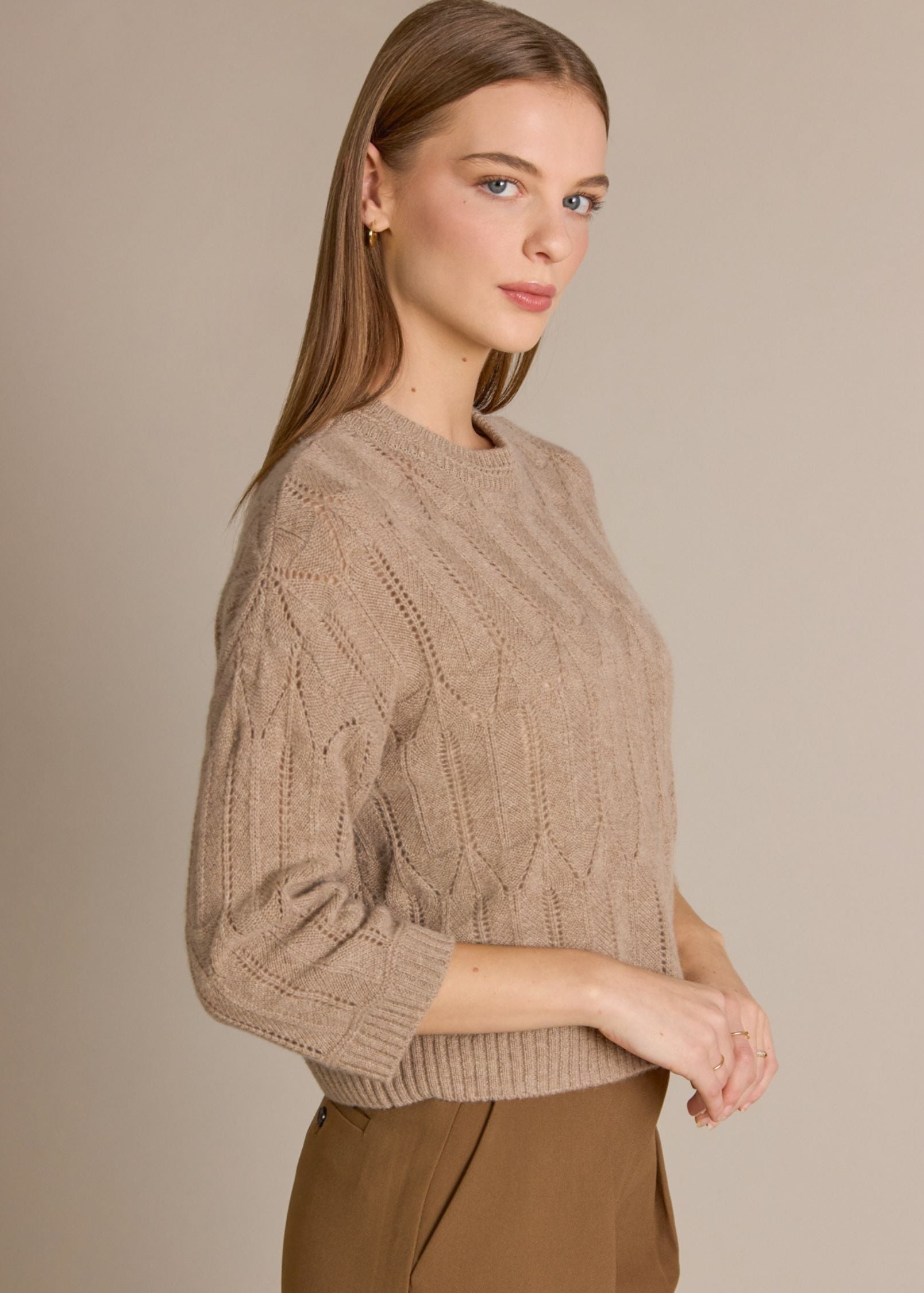 A woman wearing a beige cable-knit crewneck sweater and brown trousers, photographed against a solid warm beige studio background.