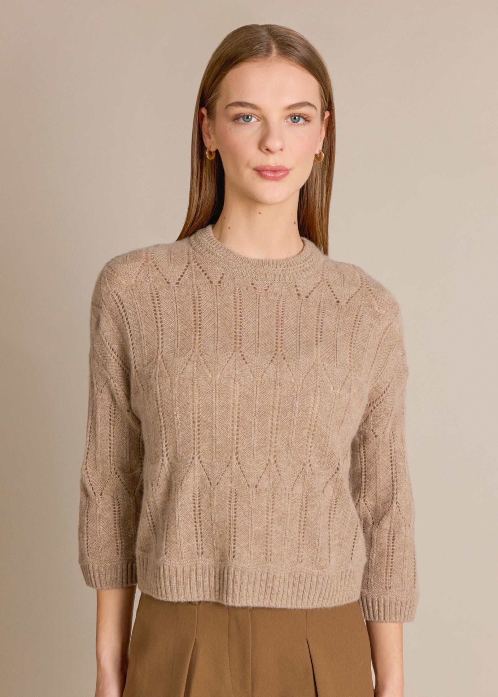 A woman modeling a beige cable-knit crewneck sweater with three-quarter sleeves, paired with brown trousers, against a solid warm beige background.