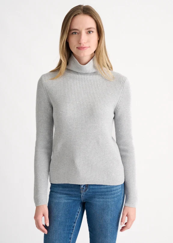 A woman wearing a light gray ribbed turtleneck sweater and blue jeans, standing against a plain white studio background.