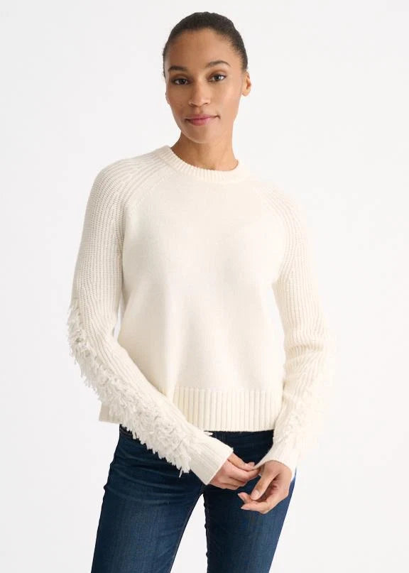 A woman wearing a cream-colored knit sweater with textured, frayed sleeves, paired with dark blue jeans, against a plain white studio background.