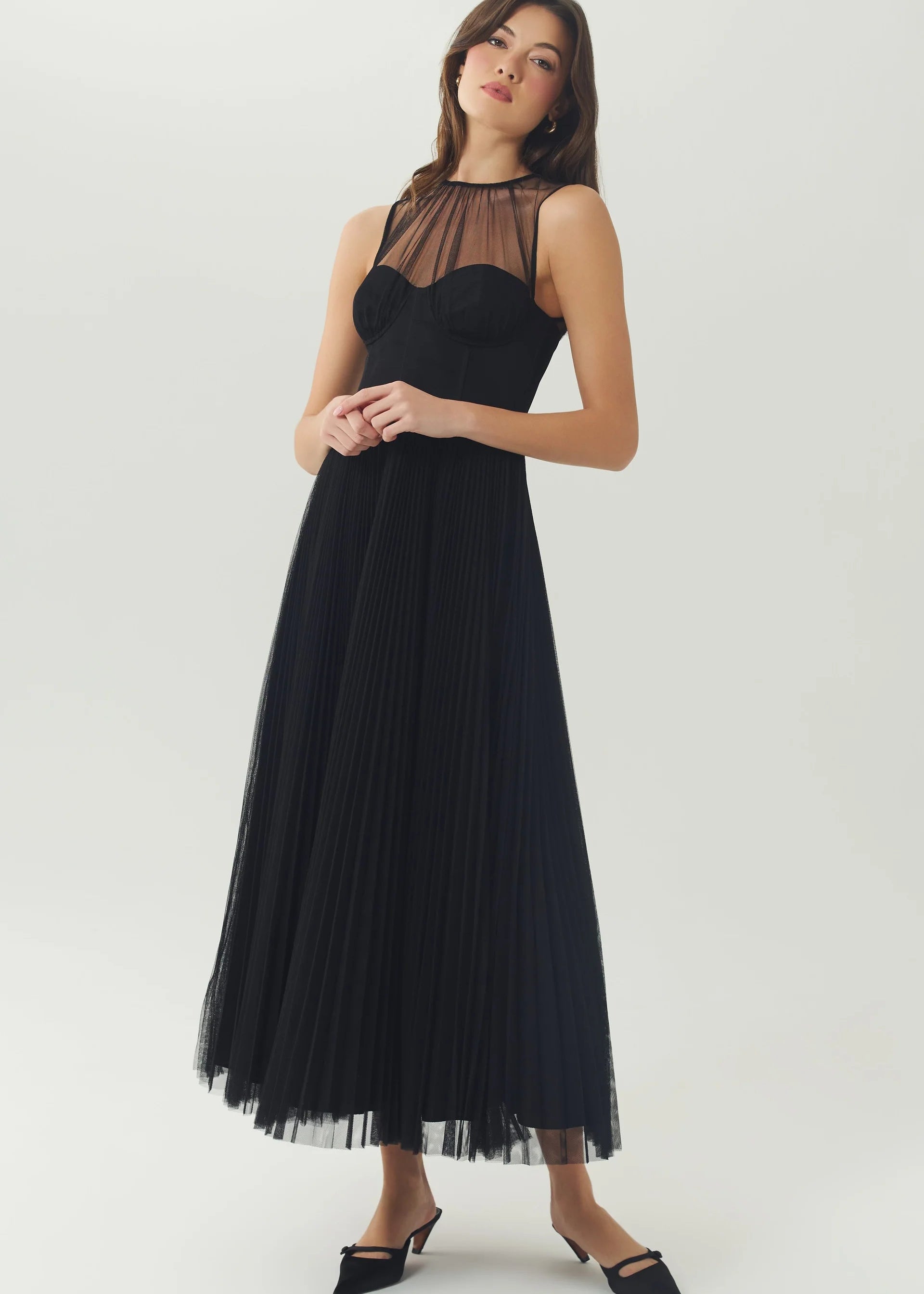 A woman modeling a sleeveless black midi dress with a sheer mesh bust and a pleated tulle skirt, paired with black pointed-toe mule heels, against a light gray studio background.