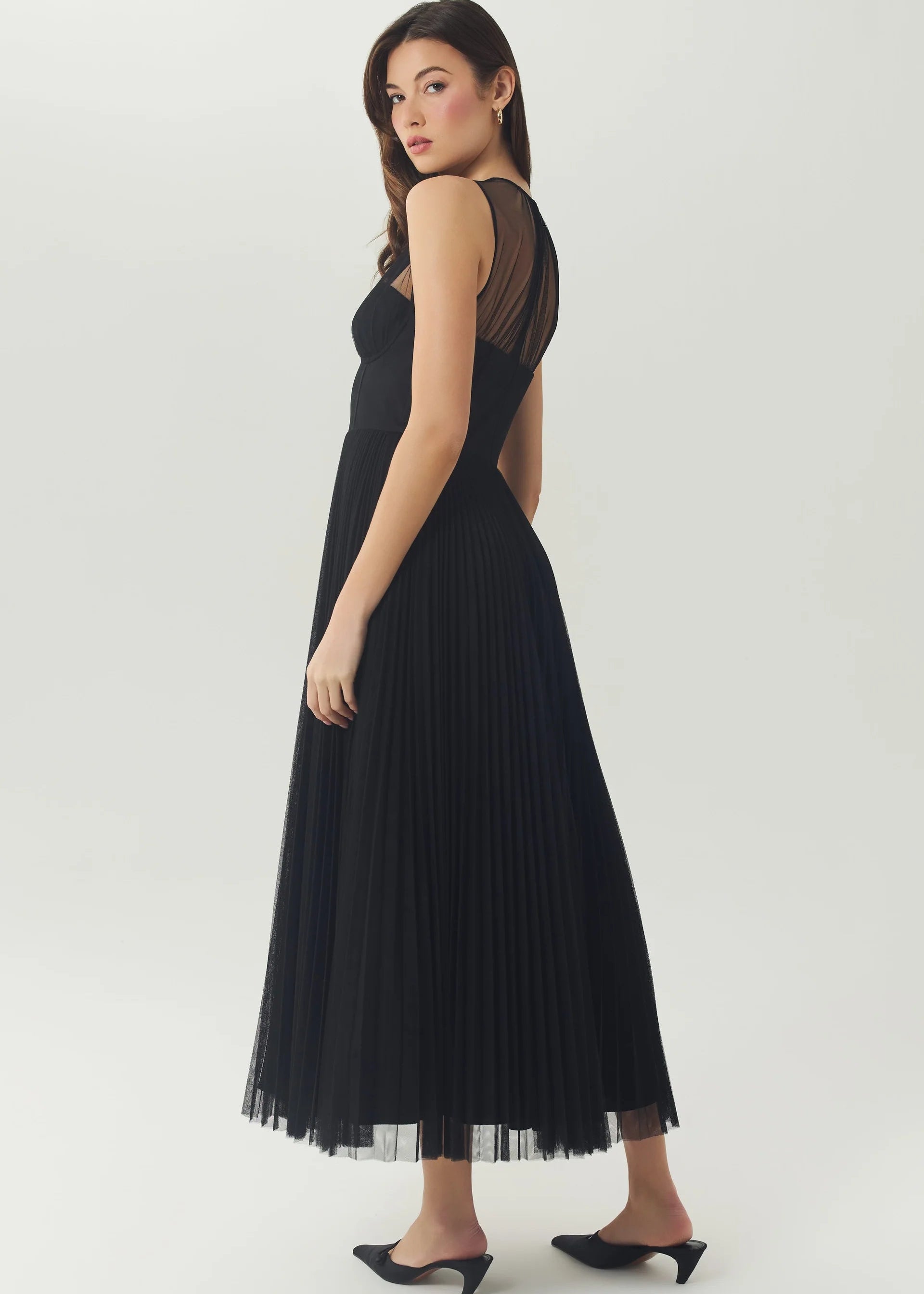 A woman modeling a sleeveless black pleated maxi dress with sheer mesh shoulder panels and side cutouts, paired with black pointed mule heels, against a light gray studio background.