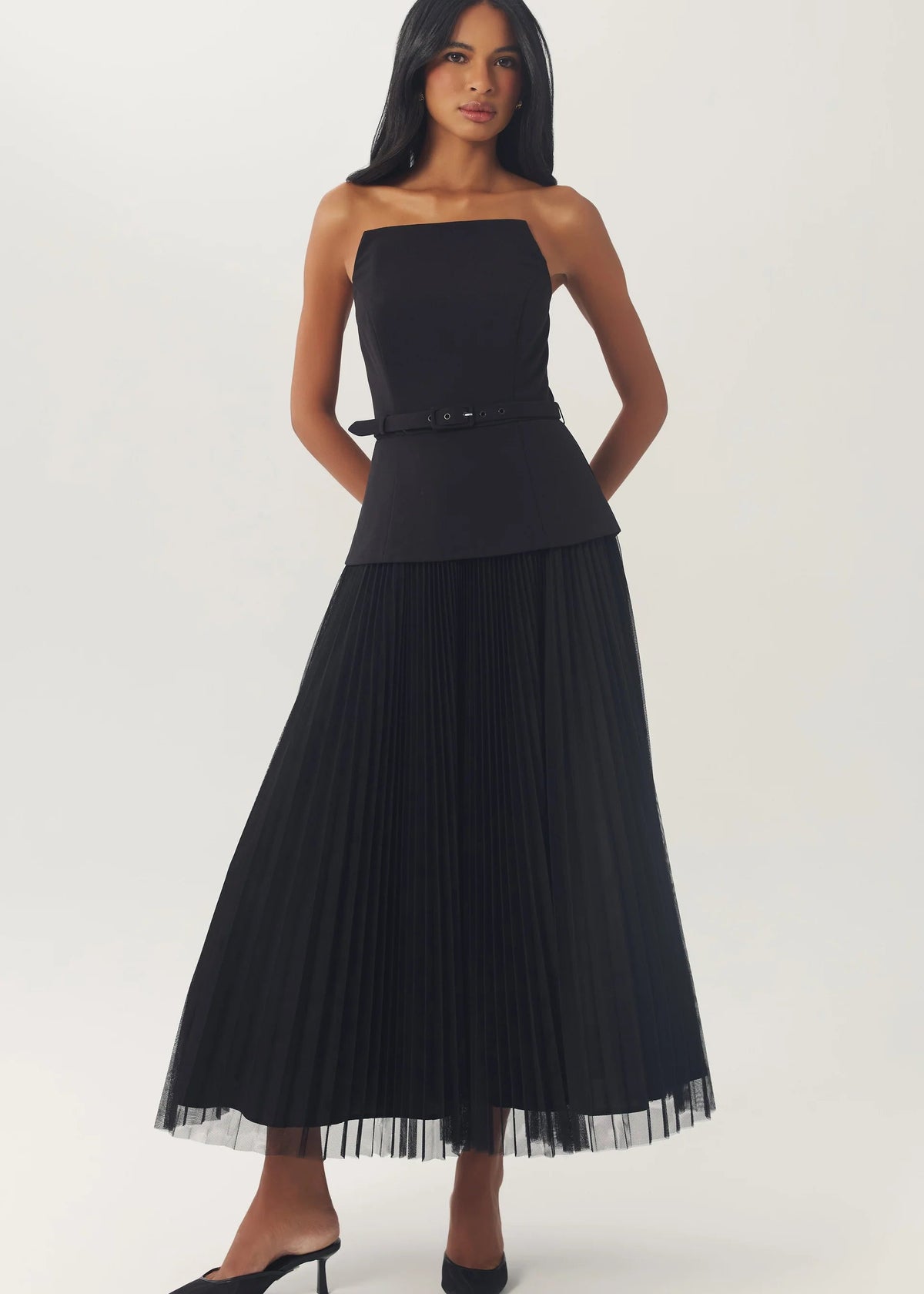 A woman modeling a black strapless pleated maxi dress with a structured bodice, thin waist tie, and sheer tulle hem, paired with black pointed-toe heels, against a plain light gray studio background.