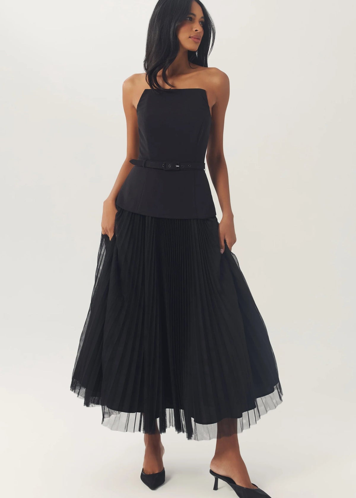 A woman modeling a black strapless midi dress with a structured bodice, thin waist belt, and a voluminous pleated tulle skirt, paired with black pointed-toe heels, against a plain light gray studio background.