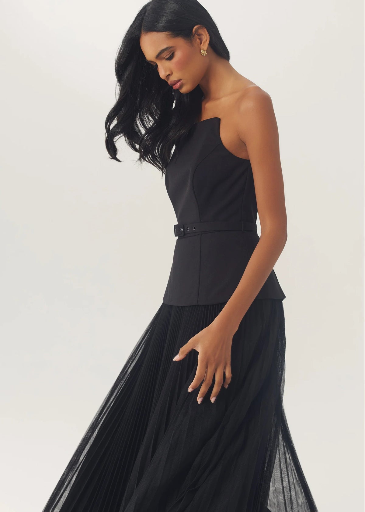 A woman modeling a black strapless gown with a structured peplum waist and a flowing sheer tulle skirt, photographed against a plain light gray studio background.