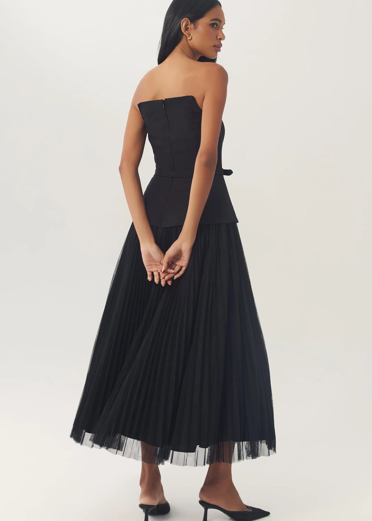 A woman modeling a strapless black midi dress with a structured bodice, peplum waist, and a full pleated skirt with a sheer tulle hem, paired with black pointed-toe heels against a plain light gray studio background.