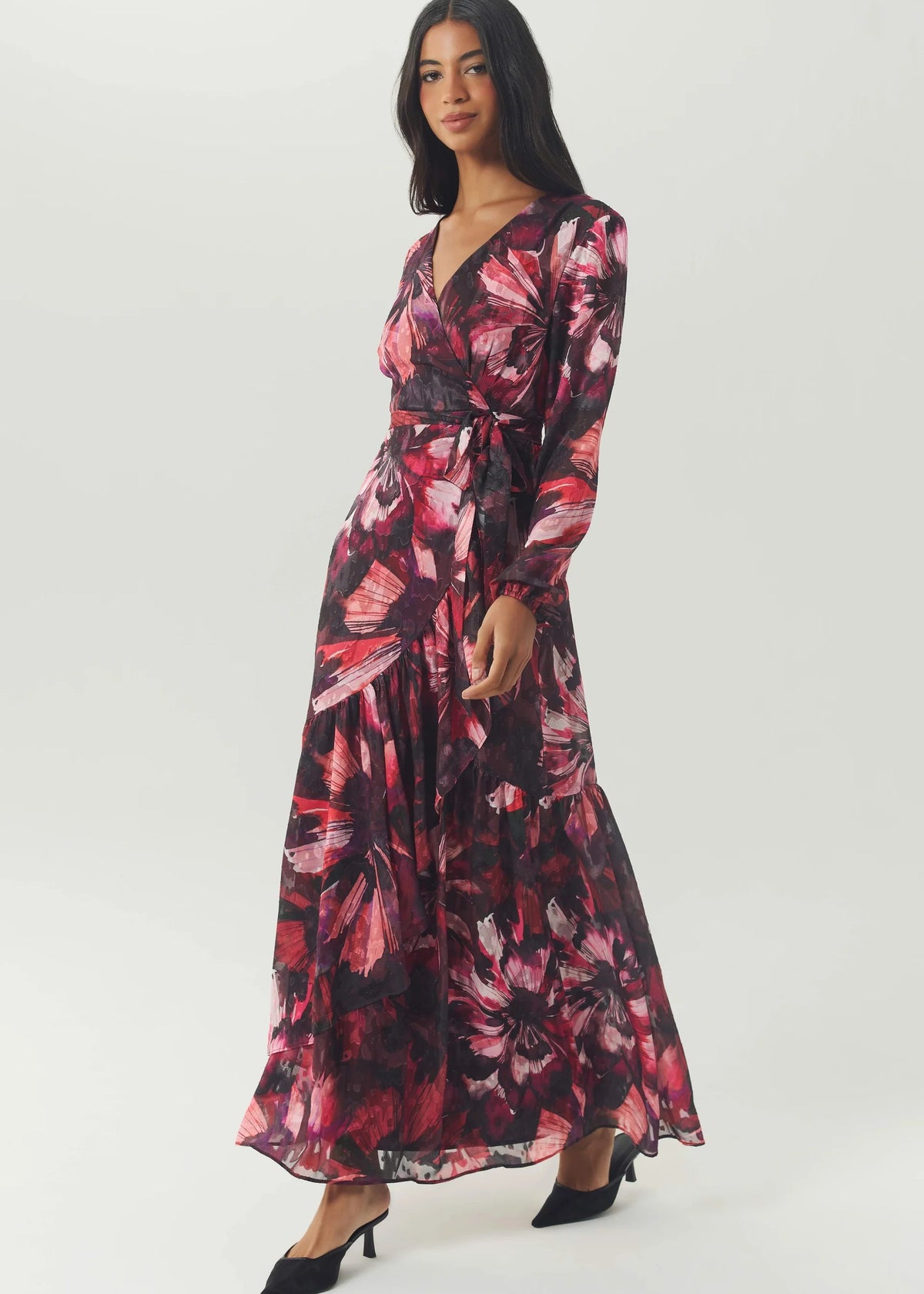 A woman modeling a long-sleeve maxi dress with a dark floral print in pink, red, and black, paired with black pointed-toe heels, against a light gray studio background.