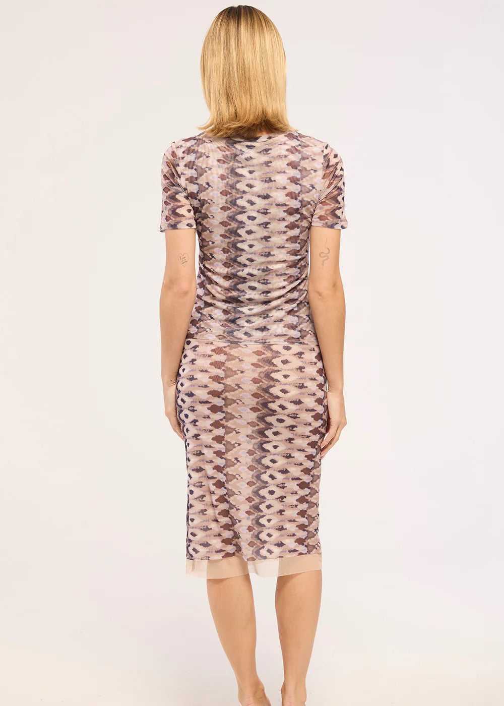 A woman viewed from the back wearing a short-sleeve bodycon dress with a pink, beige, and brown geometric zigzag pattern, paired with beige flat sandals, against a plain light background.