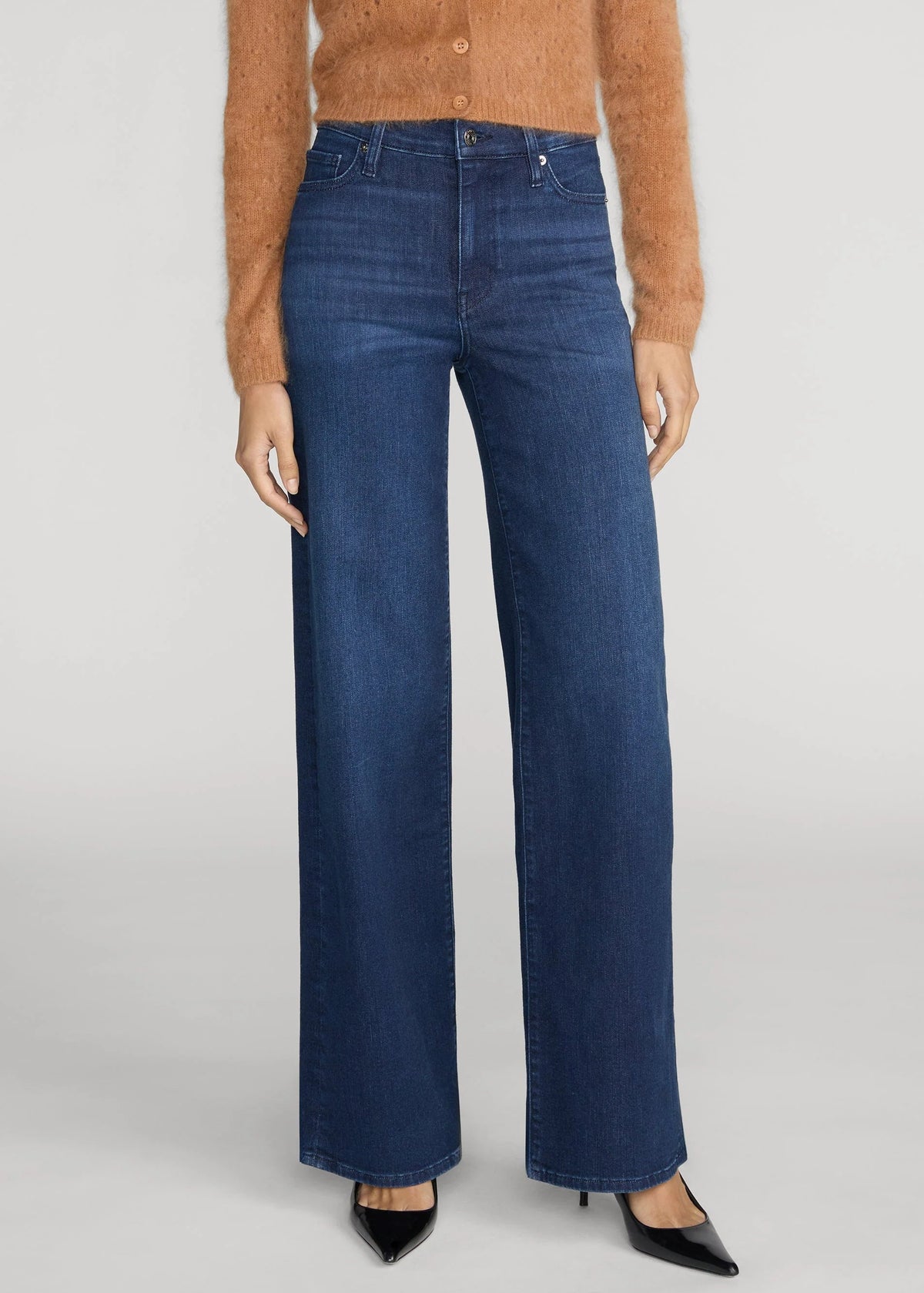 A model wearing dark blue wide-leg denim jeans, styled with a brown cardigan and black pointed-toe heels, photographed against a neutral light gray studio background.