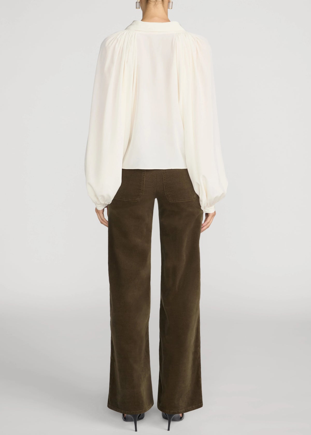 A model shown from the back wearing a white long-sleeve blouse with dramatic puffed sleeves and dark brown wide-leg trousers, set against a plain light gray studio background.