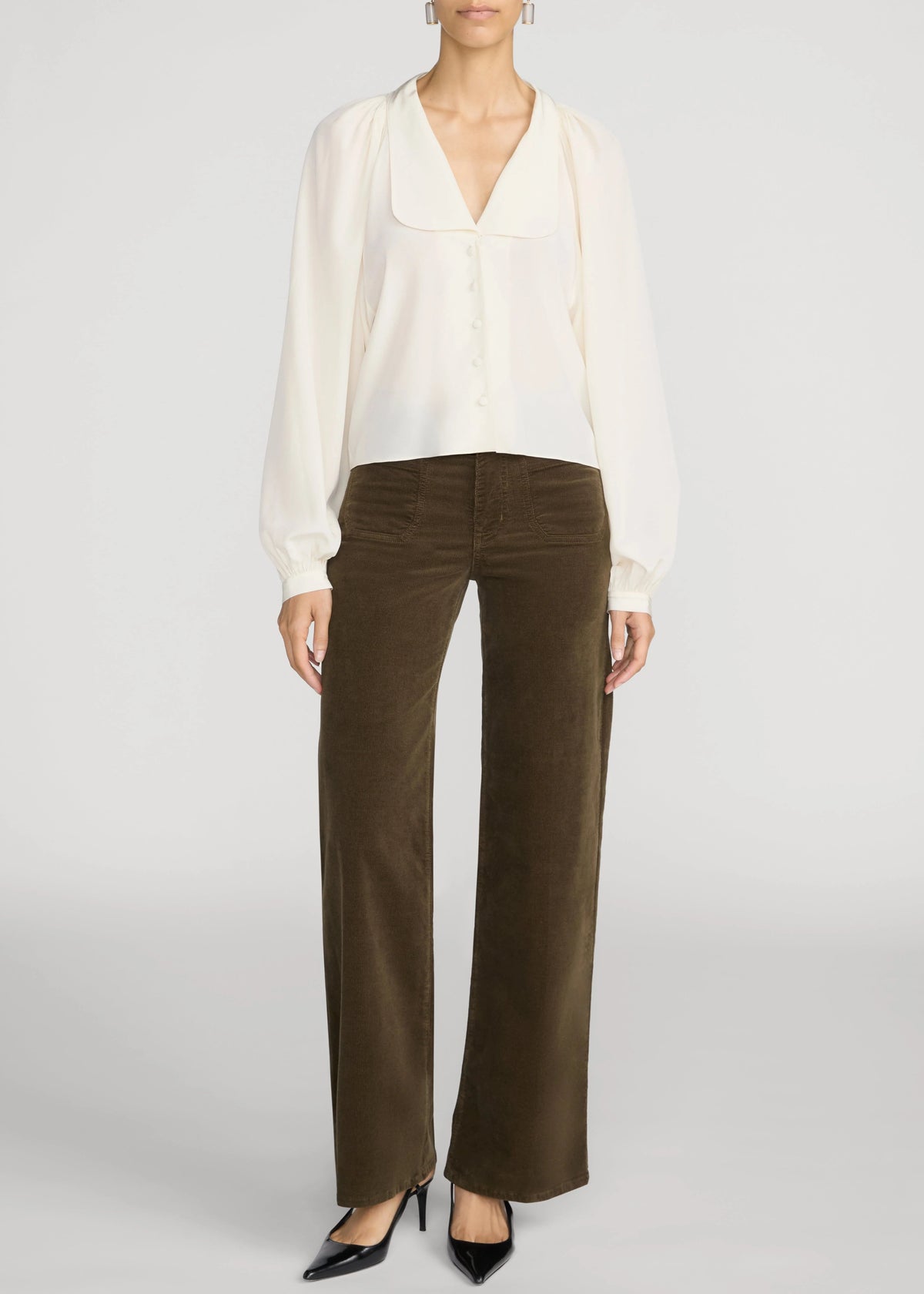 A model wearing a white long-sleeve blouse and dark brown wide-leg corduroy pants, paired with black pointed-toe heels, against a light gray studio background.