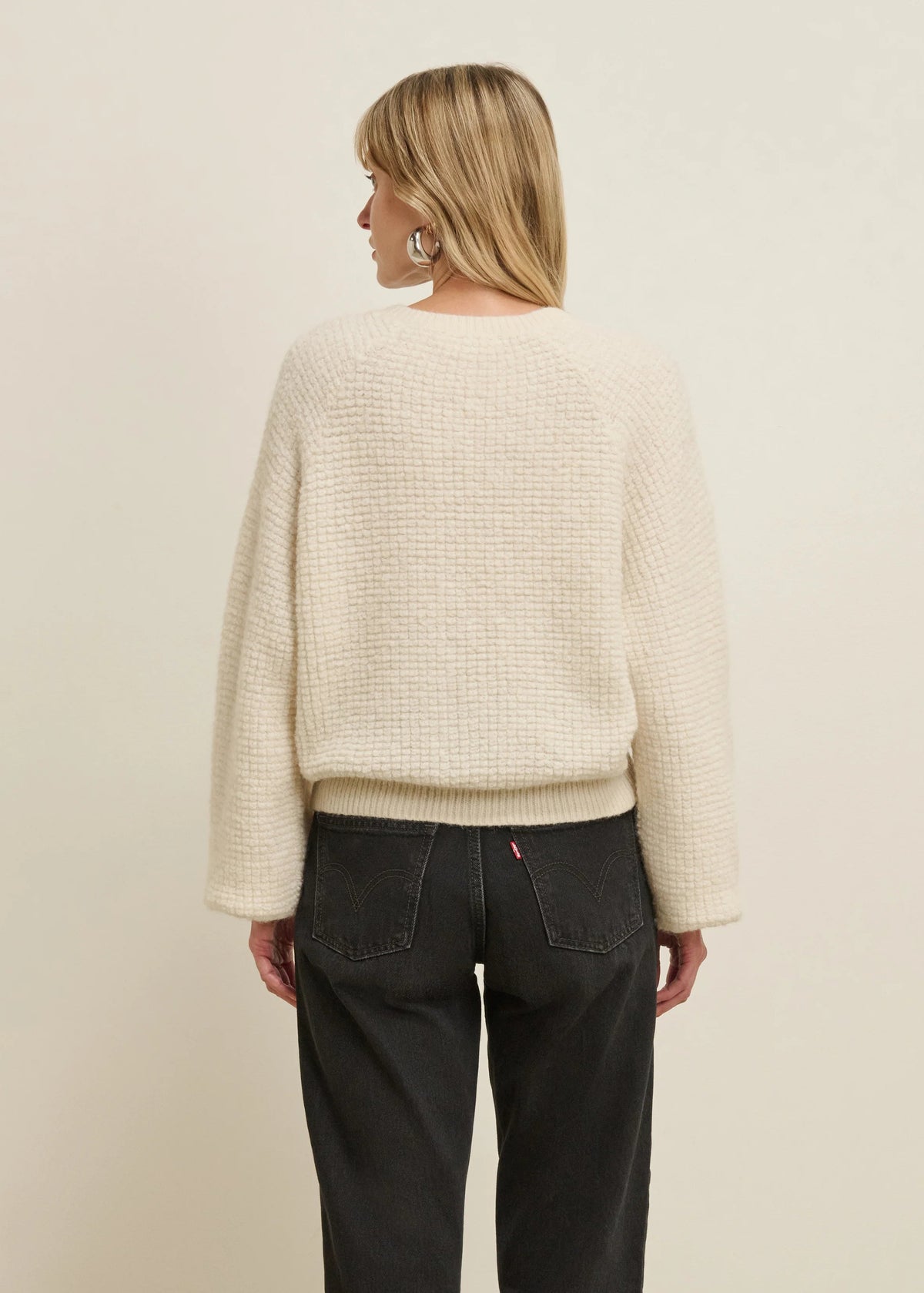 A woman shown from the back wearing a textured cream-colored knit sweater and dark charcoal gray jeans against a plain beige studio background.