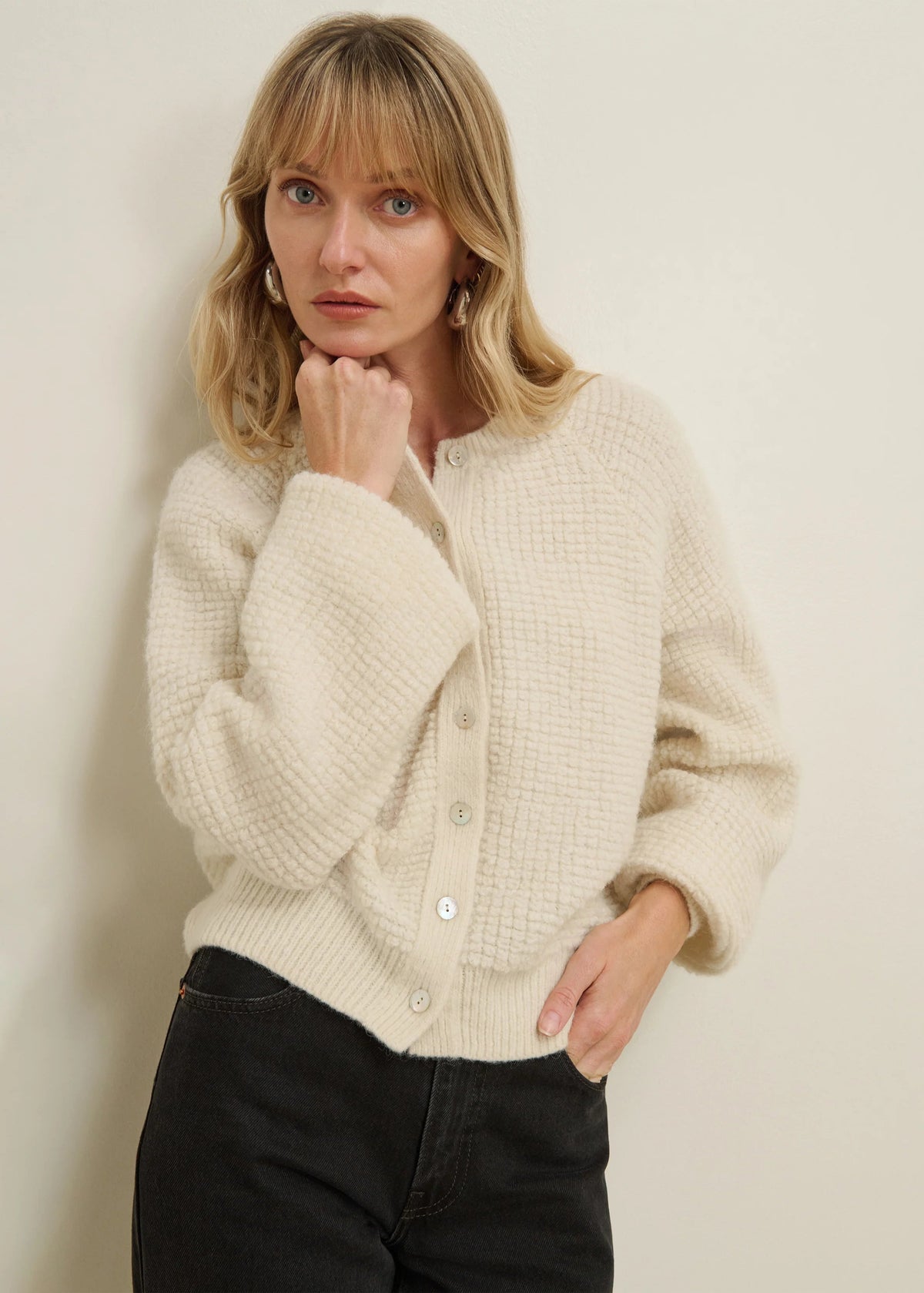 A woman with blonde hair wearing a cream-colored textured knit cardigan and black jeans, posing against a plain off-white wall.