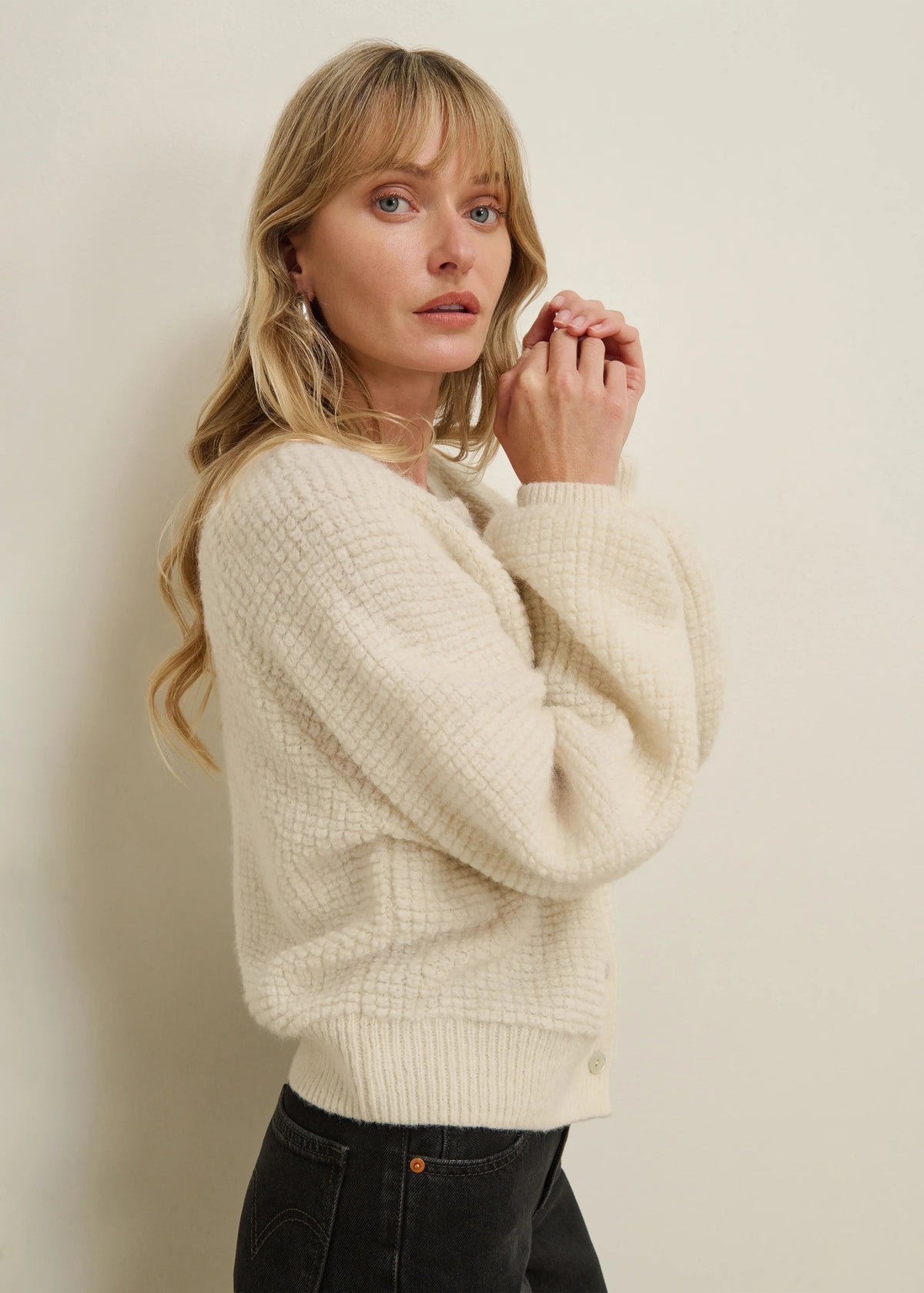 A woman with blonde hair wearing a cream-colored textured knit sweater and black jeans, posing against a plain off-white wall.