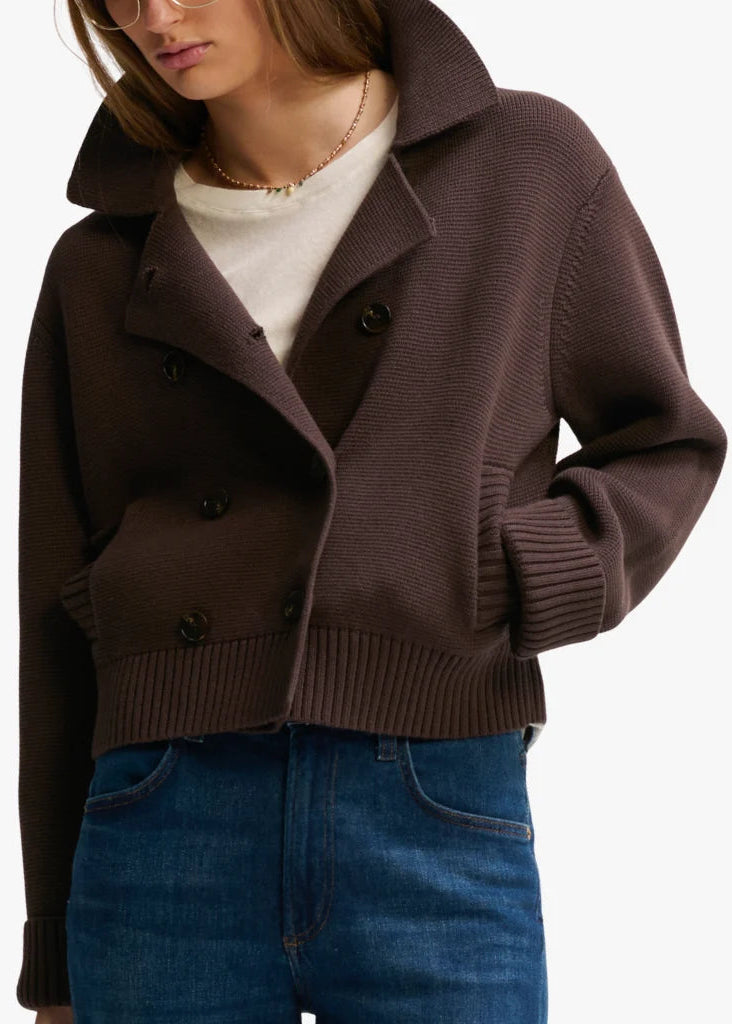 A woman wearing a dark brown ribbed knit cropped cardigan with a high collar and double-breasted buttons, layered over a cream turtleneck and blue jeans.