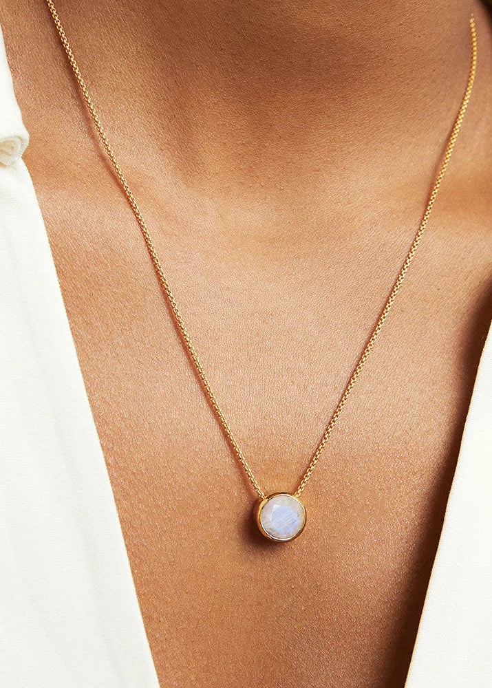 A close-up of a person wearing a delicate gold chain necklace with a round white opal pendant, styled with an open white shirt.
