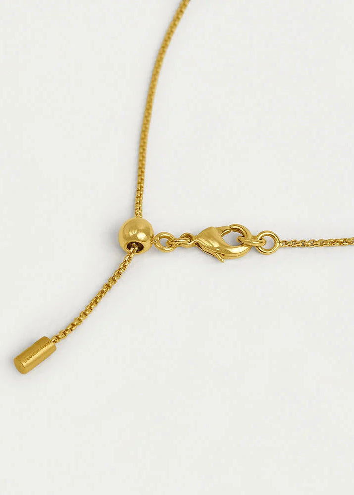 Close-up of a gold chain necklace with an adjustable lobster clasp, a small bead, and a dangling extender with a cylindrical tip, shown on a plain light background.