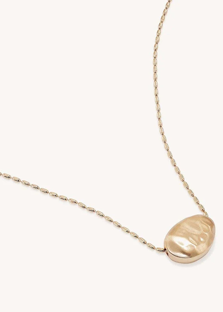 A delicate gold chain necklace featuring a large, irregularly shaped gold pendant, displayed on a clean off-white background.