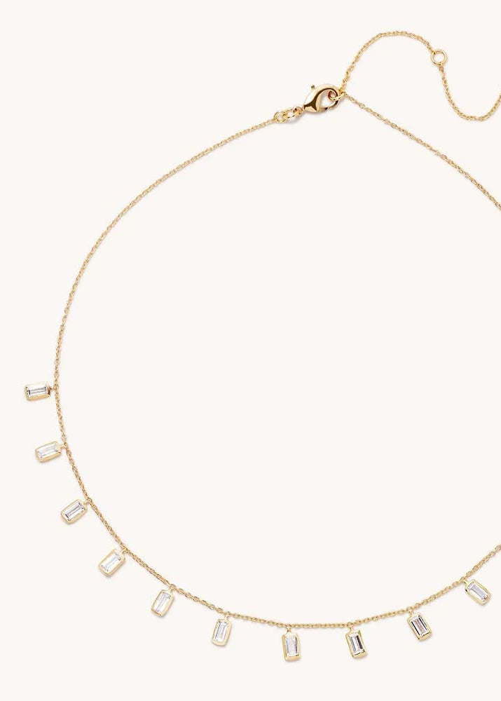 A delicate gold chain necklace with a row of small rectangular clear baguette-cut stones, displayed on a light cream background.