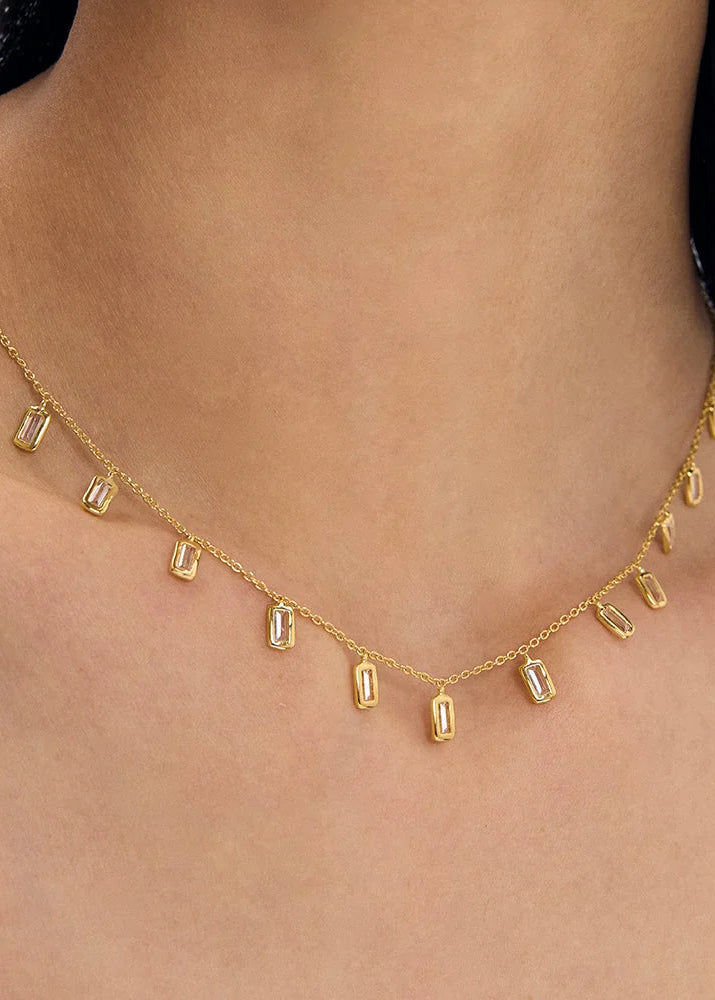A delicate gold chain necklace with small rectangular clear gemstones, shown worn on a model's neck and upper chest.