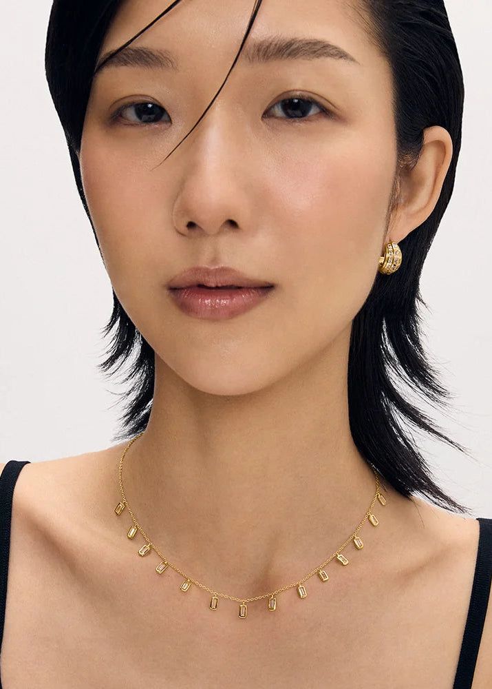 A close-up portrait of a woman wearing a delicate gold necklace with small dangling leaf-like charms and a matching gold stud earring, against a clean light gray background.