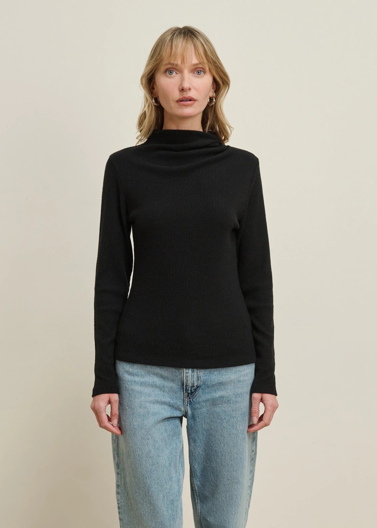 A woman modeling a black long-sleeve cowl-neck sweater paired with light-wash blue jeans, standing against a plain beige studio background.