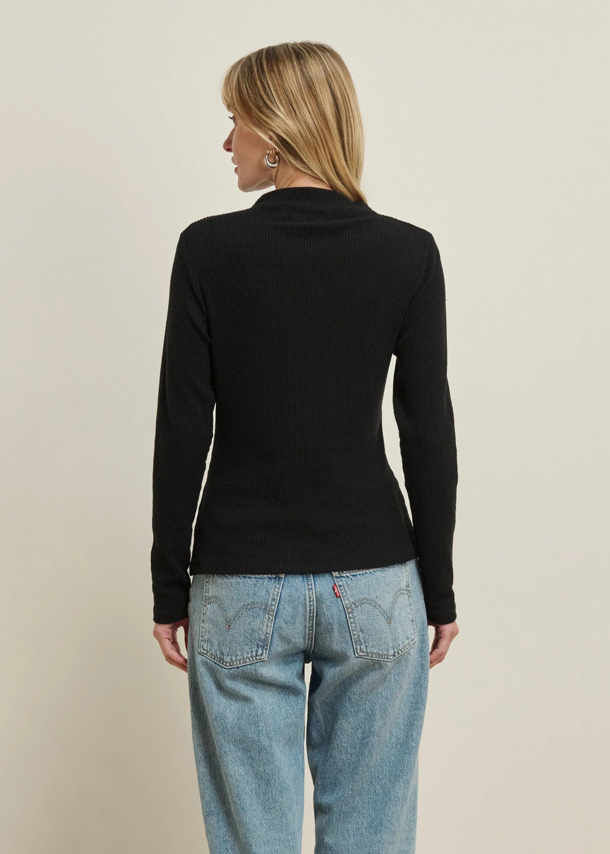 A woman viewed from the back wearing a fitted black long-sleeve mock neck top and light blue jeans against a plain beige studio background.