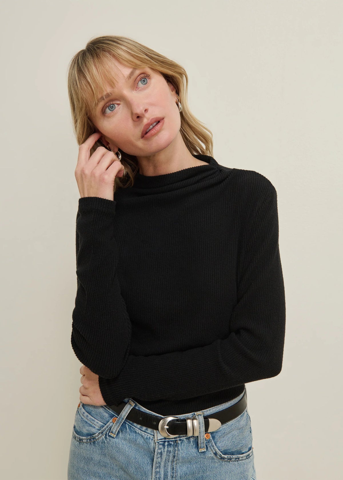 A woman wearing a black ribbed mock neck long-sleeve top and light wash blue jeans with a black belt, posing against a neutral beige background.