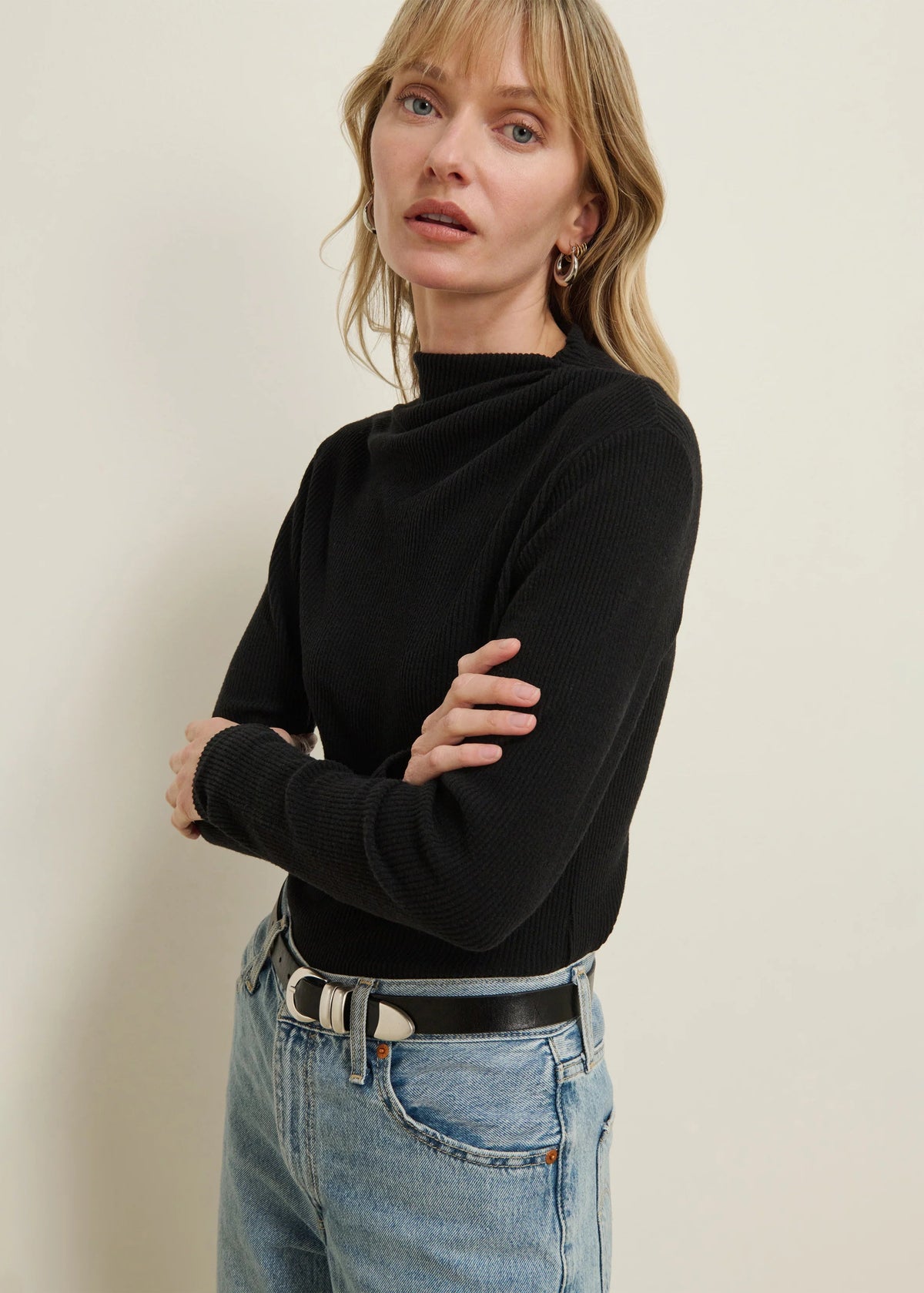 A woman wearing a black ribbed mock neck long-sleeve sweater and light wash blue jeans with a black belt, standing against a plain beige wall.