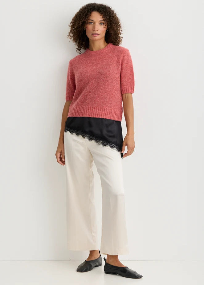 A model stands against a plain white studio background wearing a short-sleeve pink knit sweater, a black satin underlayer with lace trim, off-white wide-leg cropped pants, and black flats.
