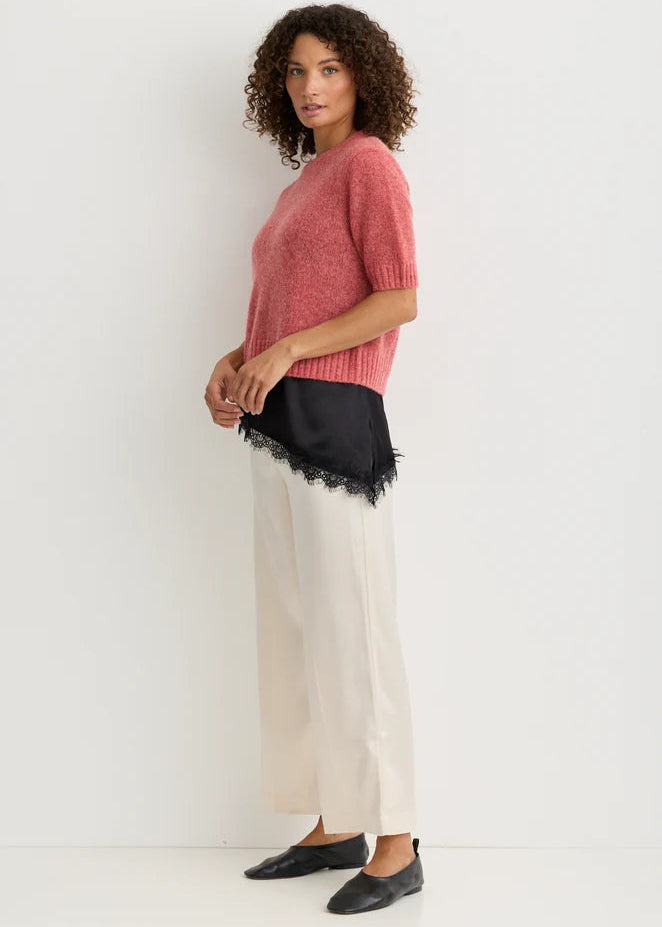 A woman modeling a short-sleeve pink knit sweater paired with a black satin waistband featuring a tassel and a black lace fringe, styled with wide-leg cream trousers and black flats against a white studio background.