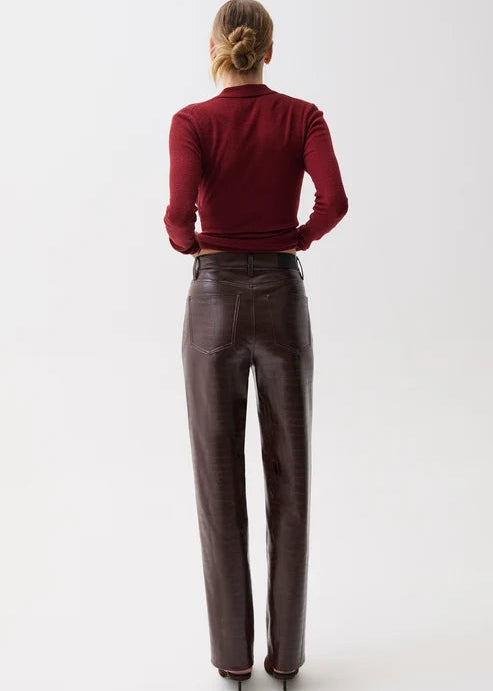 A woman viewed from behind wearing a dark red long-sleeve mock-neck top and dark brown leather trousers, standing against a plain white studio background.