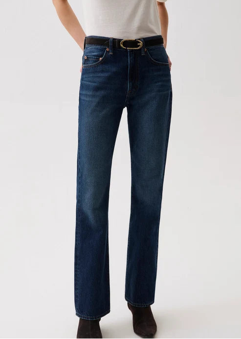 A model wearing dark blue straight-leg denim jeans with a black leather belt featuring a gold-tone oval buckle, styled with a white t-shirt and dark brown boots against a plain white studio background.