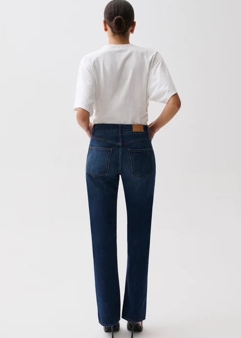 A model viewed from behind wearing dark blue straight-leg jeans, a white t-shirt, and black heels against a plain white studio background.