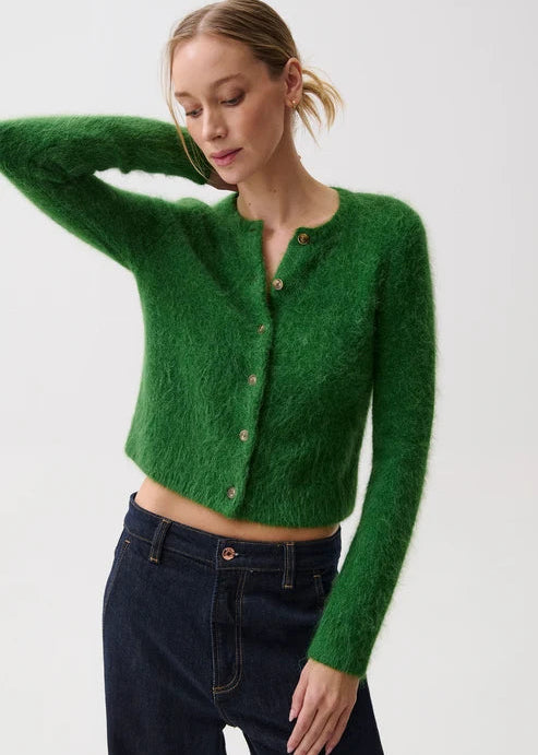 A woman modeling a fuzzy, cropped green button-up cardigan sweater paired with dark blue jeans against a plain white studio background.