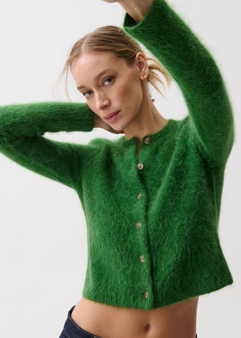 A woman wearing a fuzzy green cropped button-up cardigan sweater and dark blue jeans, posing against a light gray studio background.