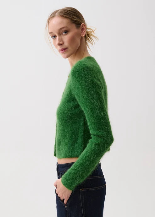 A woman wearing a fuzzy green cropped long-sleeve sweater and dark blue jeans, photographed against a plain light gray studio background.