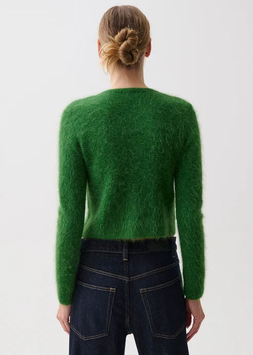 A woman viewed from the back wearing a bright green fuzzy long-sleeve sweater and dark blue jeans against a plain light gray studio background.