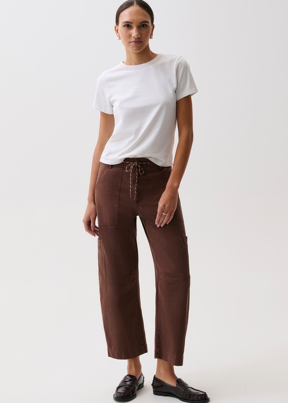 A model stands against a plain light background wearing a white short-sleeve crewneck t-shirt, brown wide-leg cropped pants with a drawstring waist, and black loafers.