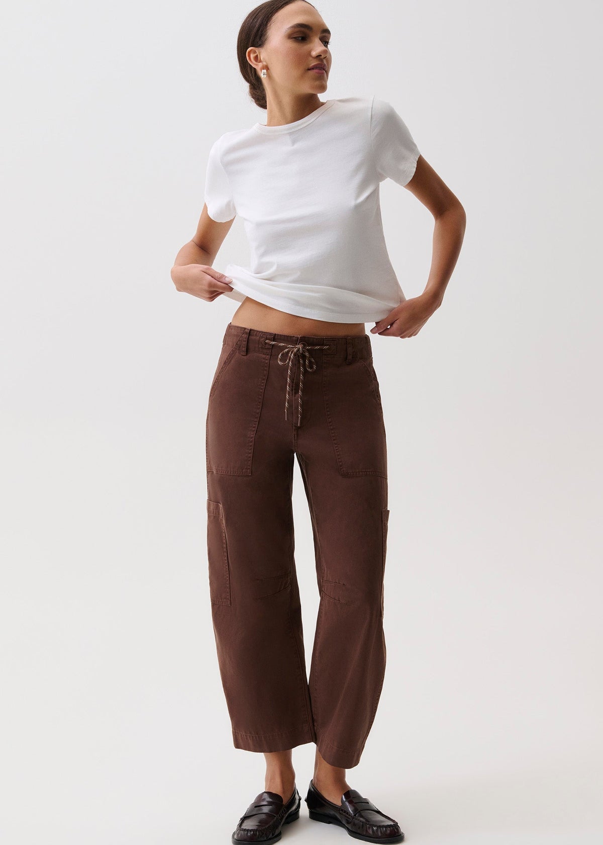 A woman modeling a white short-sleeve t-shirt and dark brown wide-leg cropped pants with a drawstring waist, paired with black loafers, against a plain light gray studio background.