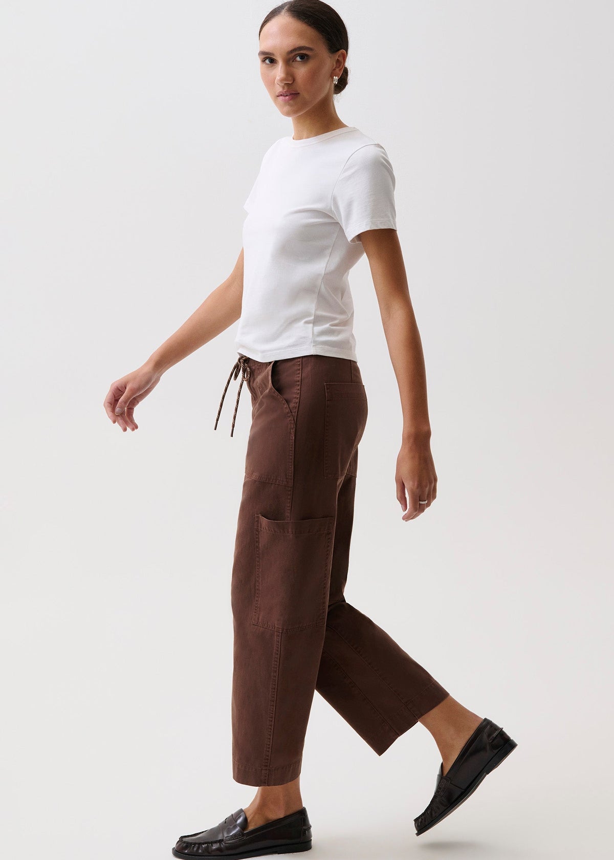 A woman modeling a pair of dark brown cropped trousers with a drawstring waist, styled with a white t-shirt and black loafers against a plain light gray studio background.