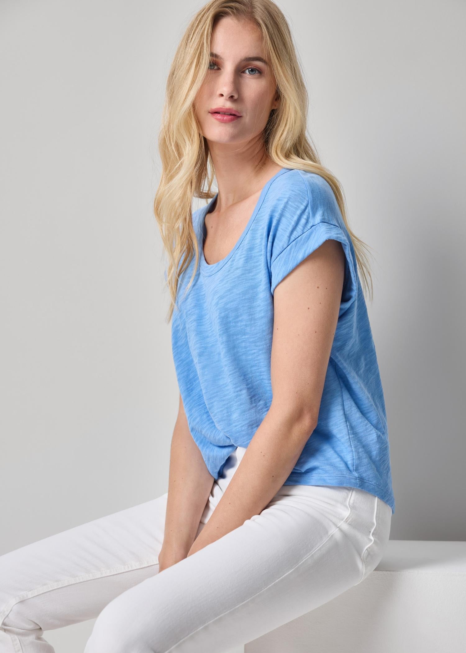 Woman wearing a blue shirt and white pants sitting on a white surface with a light gray background