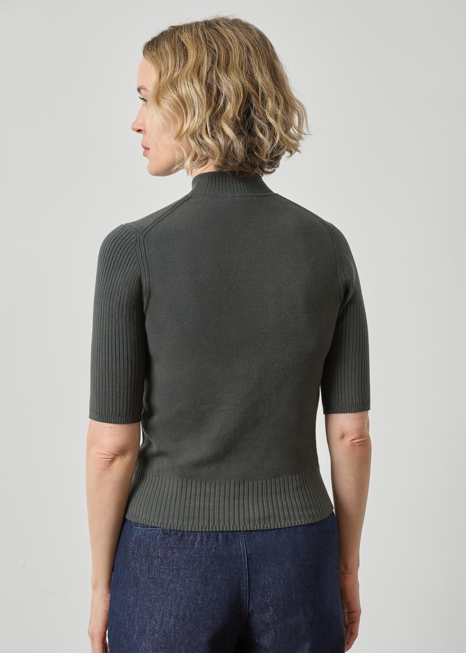 A woman shown from the back wearing a dark olive green short-sleeve ribbed knit mock-neck sweater and blue jeans against a light gray studio background.