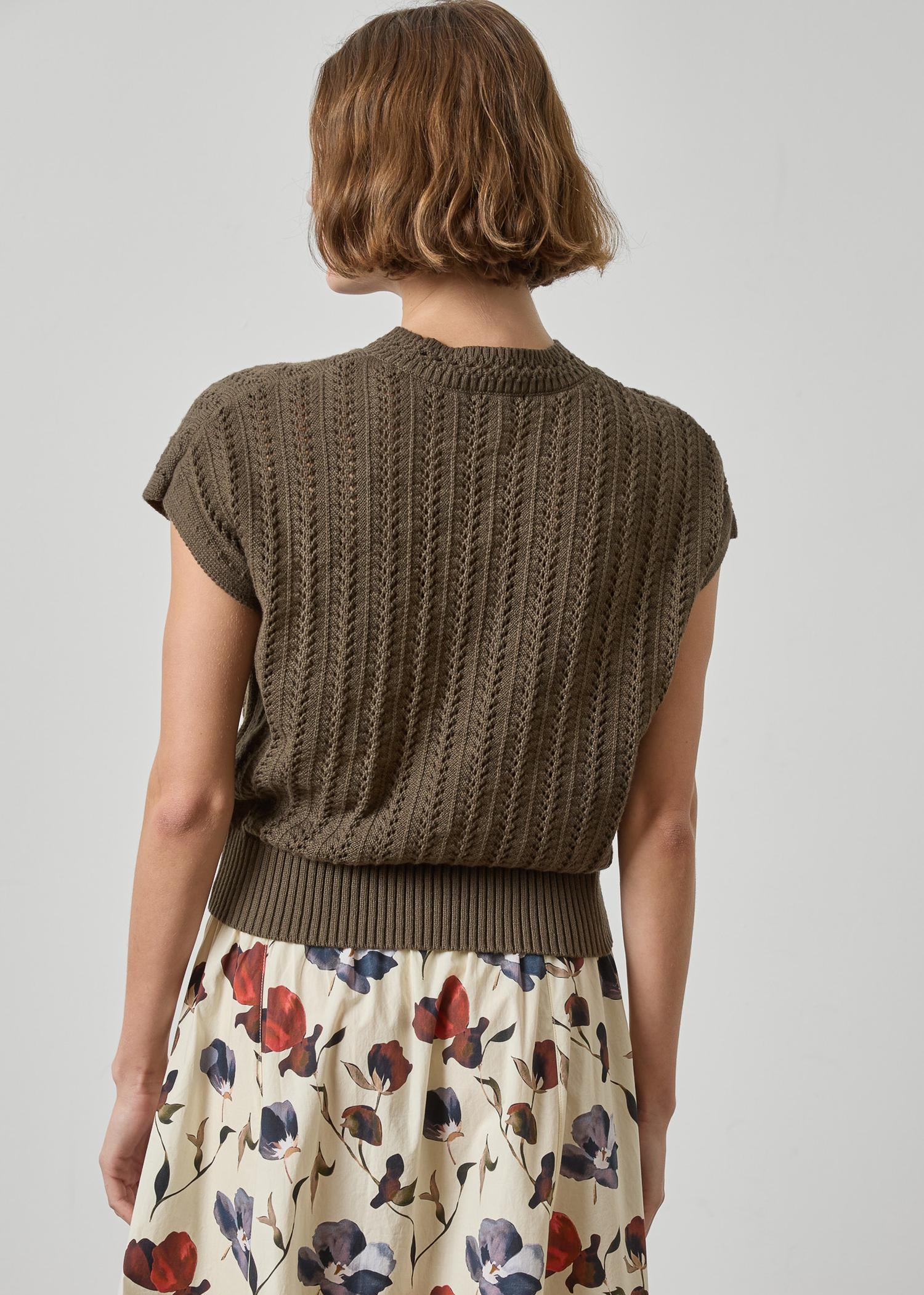 Woman wearing a brown knitted sleeveless sweater over a floral dress on a plain background