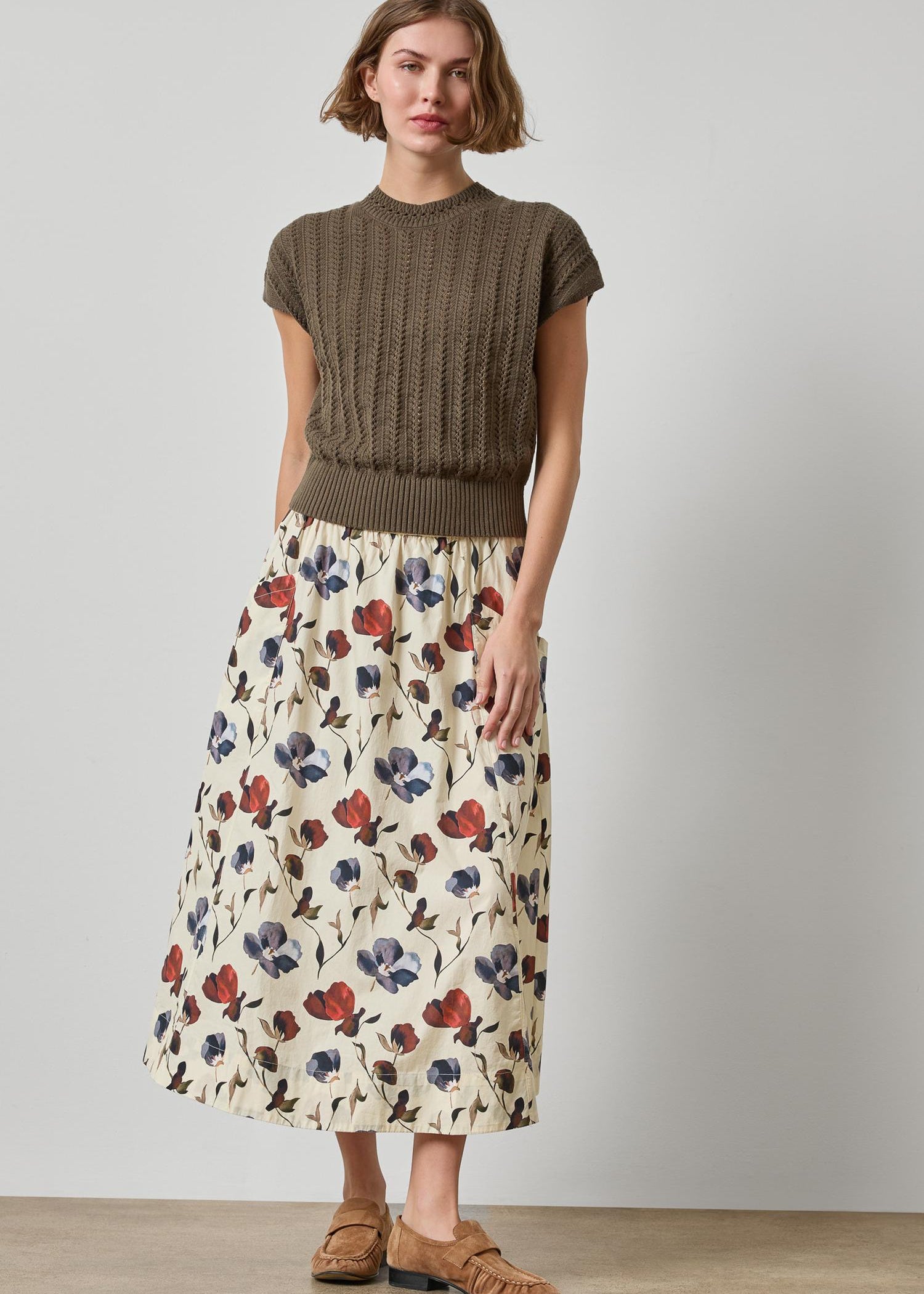 Woman wearing a brown sweater and floral skirt against a plain background