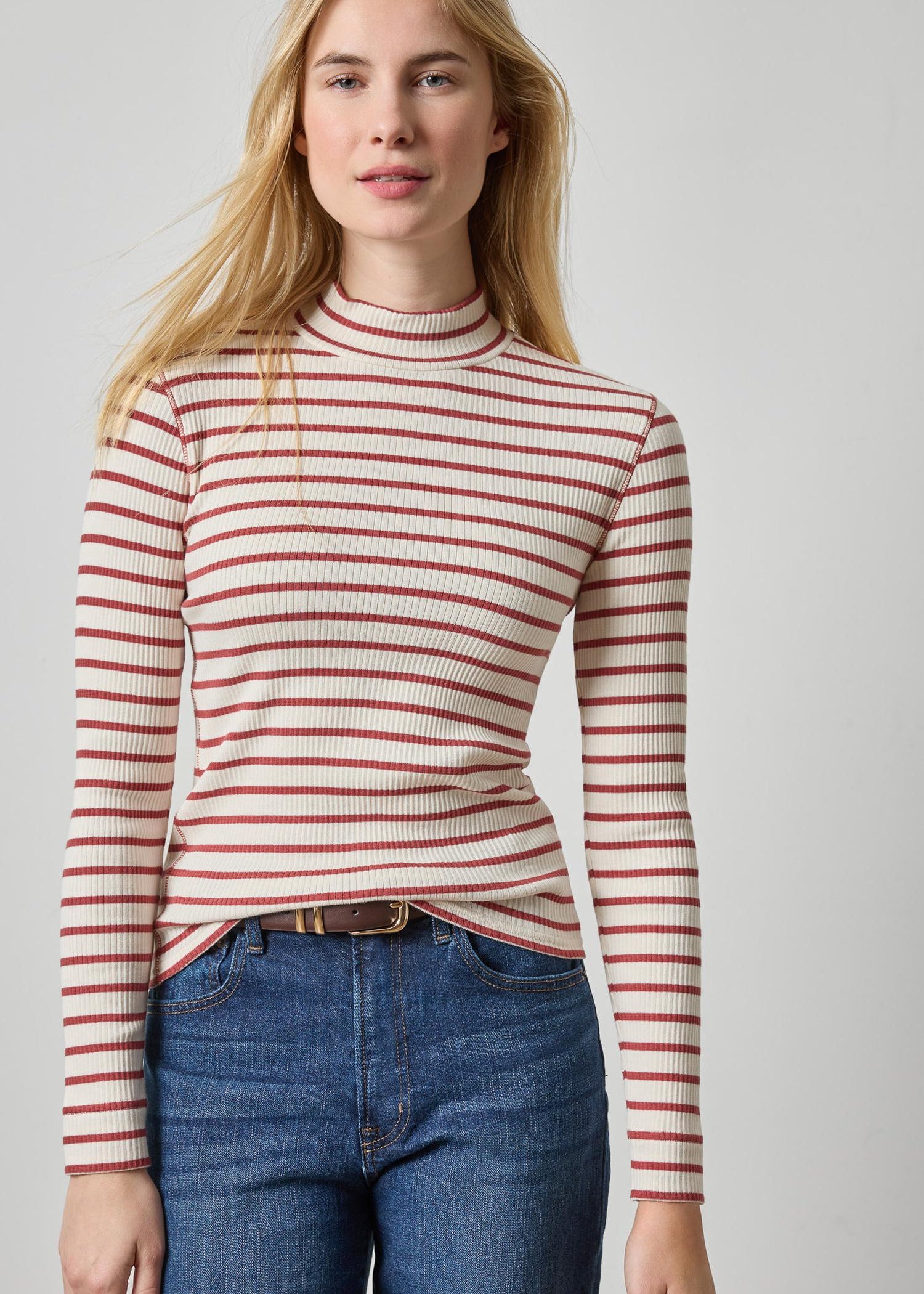 A woman wearing a fitted long-sleeve ribbed mock neck top with horizontal red stripes on a cream background, paired with blue jeans, against a light gray studio background.