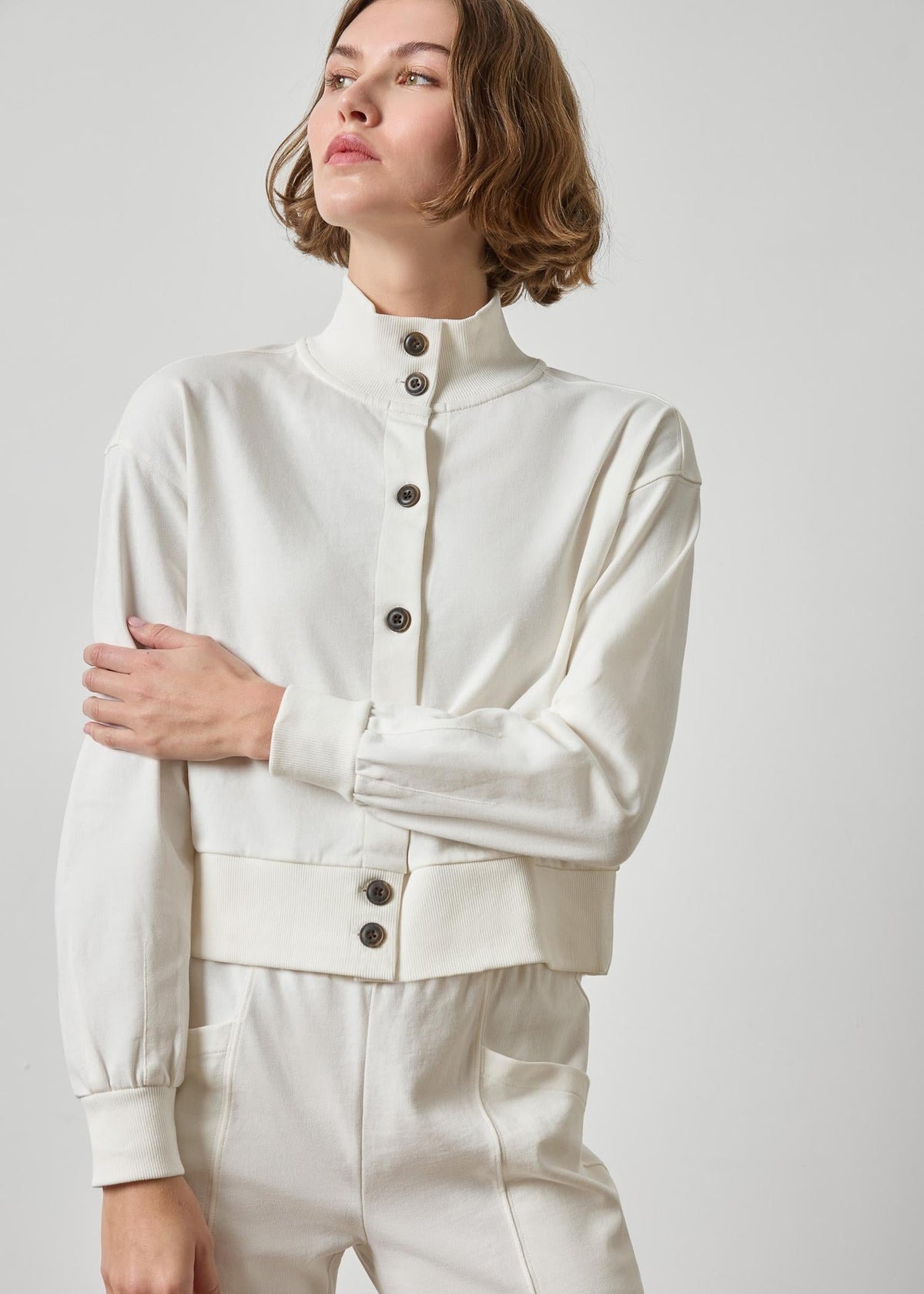 A woman wearing a cream-colored long-sleeve cropped jacket with a high mock neck and dark buttons, paired with matching cream pants, against a light gray studio background.